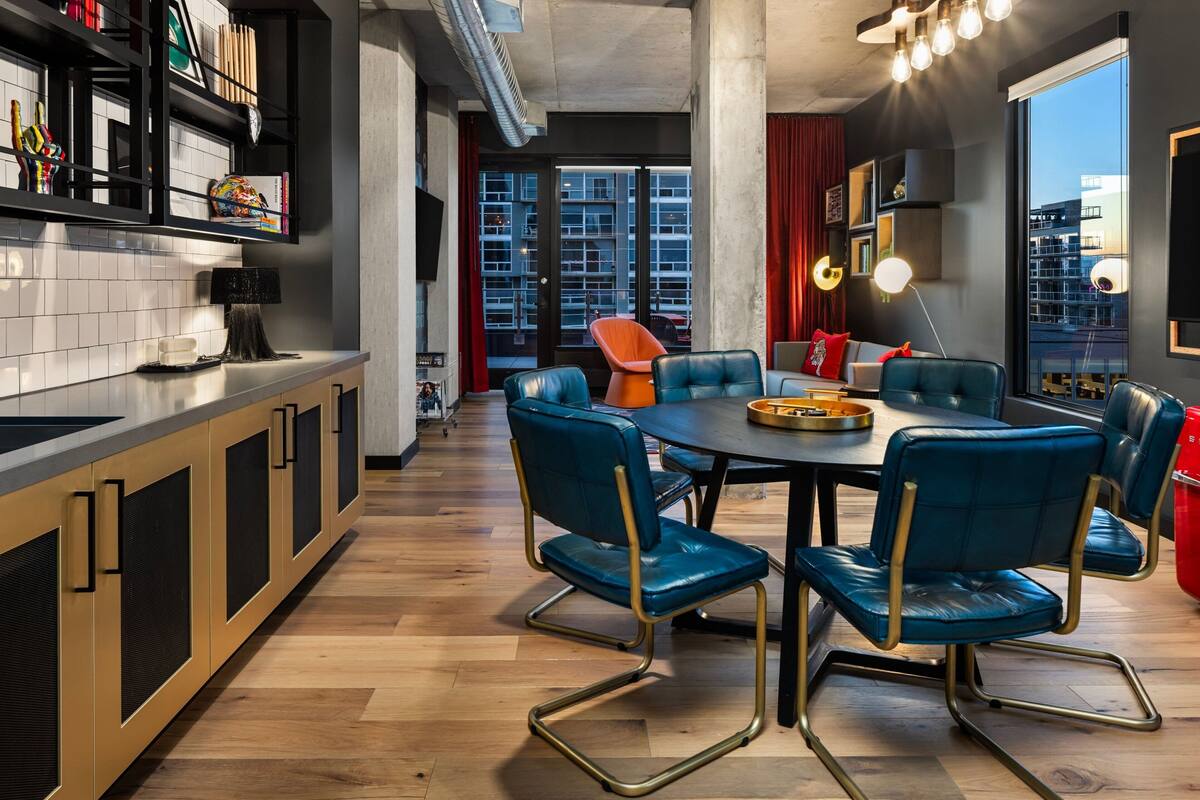 A modern kitchen with a dining area, featuring a round table with blue chairs, a black countertop, and a large window with a city view.