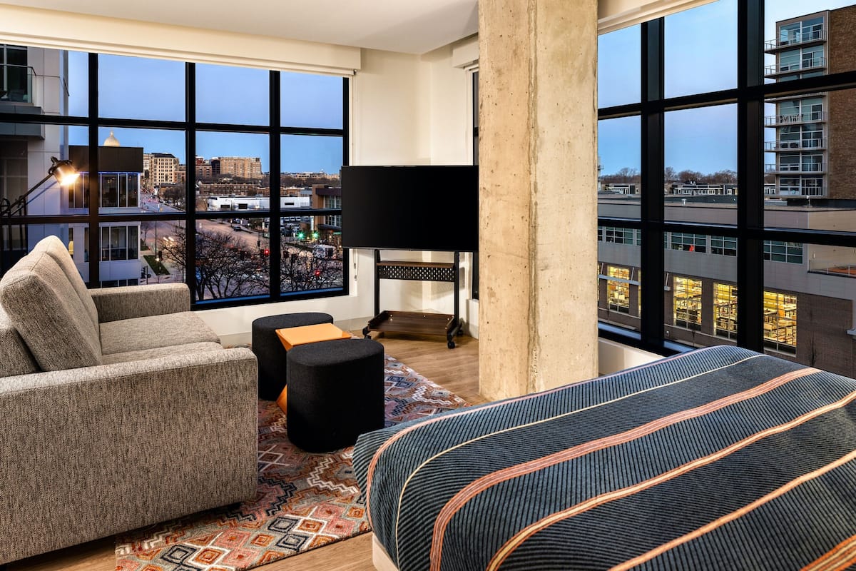 A modern hotel room with large windows, a sofa, a bed, and a lamp.