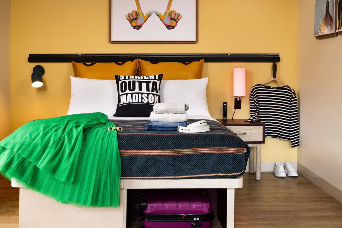 A neatly made bed with a green blanket, a pillow with "STRAIGHT OUTTA MADISON" text, and a purple suitcase under the bed.