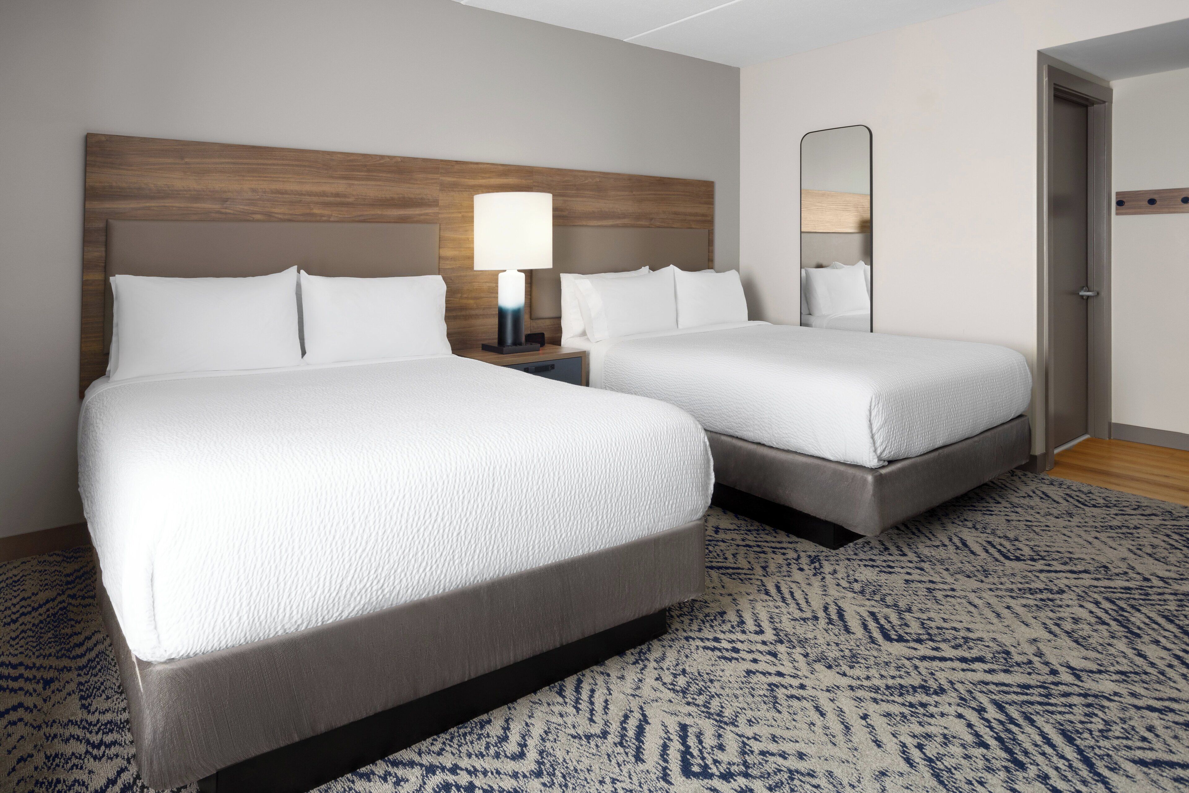 A hotel room with two beds, a nightstand with a lamp, a mirror, and a patterned carpet.
