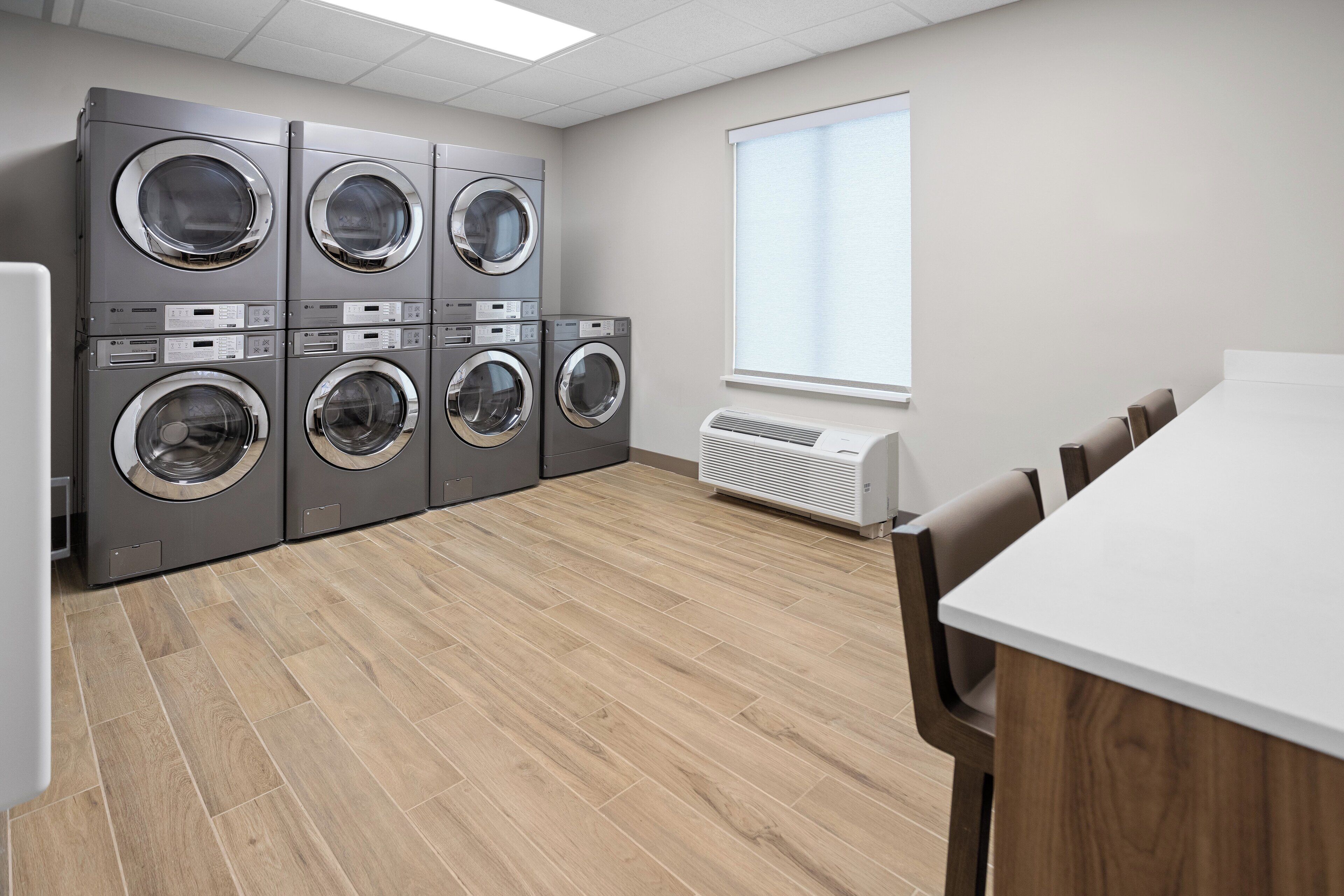 A laundry room with six stacked front-loading washers and dryers, a large window with blinds, and a white table with chairs.