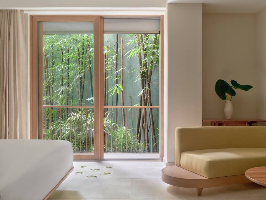 Una habitación de hotel moderna con un gran ventanal corredero que da a un jardín de bambú, un sofá y una pequeña mesa.