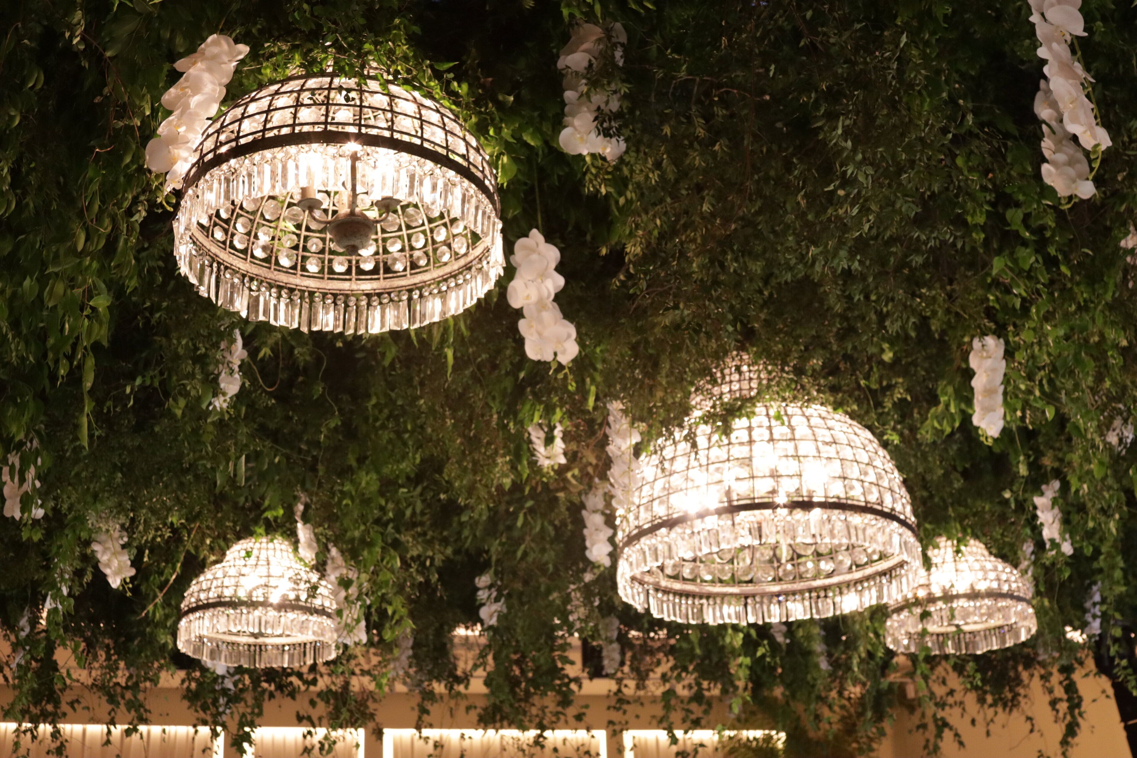A chandelier with multiple tiers of crystals hanging from a ceiling, surrounded by lush greenery and white flowers.