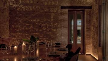 A dimly lit dining area with a rustic brick wall, a wooden table set for a meal, and a door leading to another room.