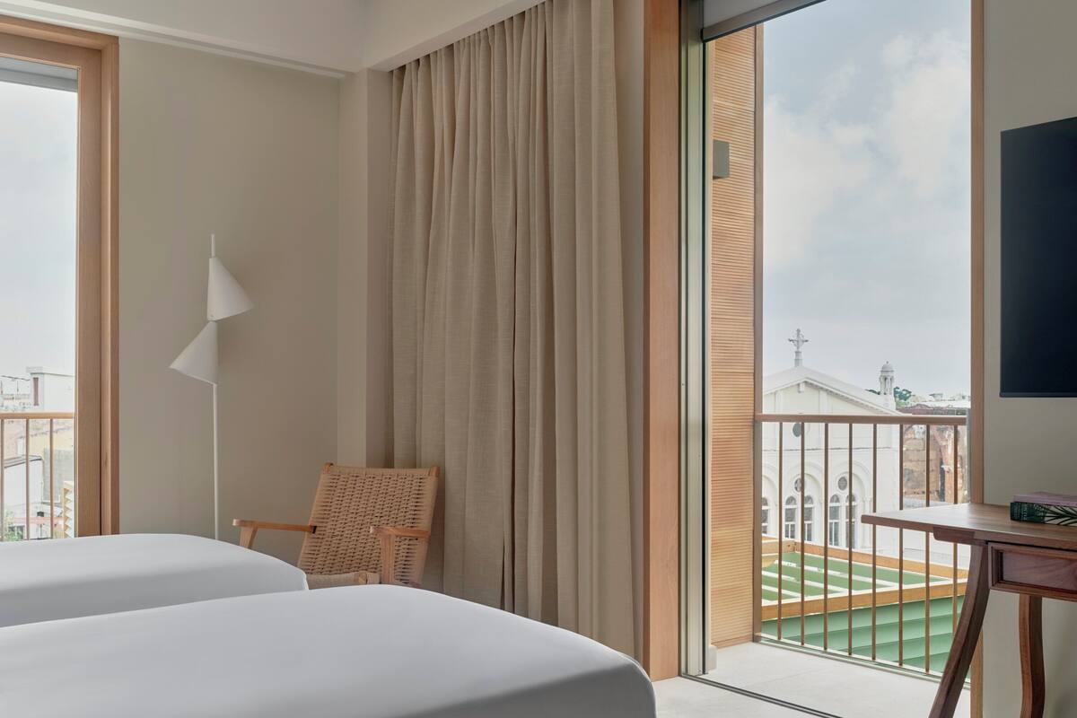 Hotellihuone, jossa on kaksi sänkyä, rottinkituoli, pieni pöytä ja näkymä rakennukseen suuresta ikkunasta.