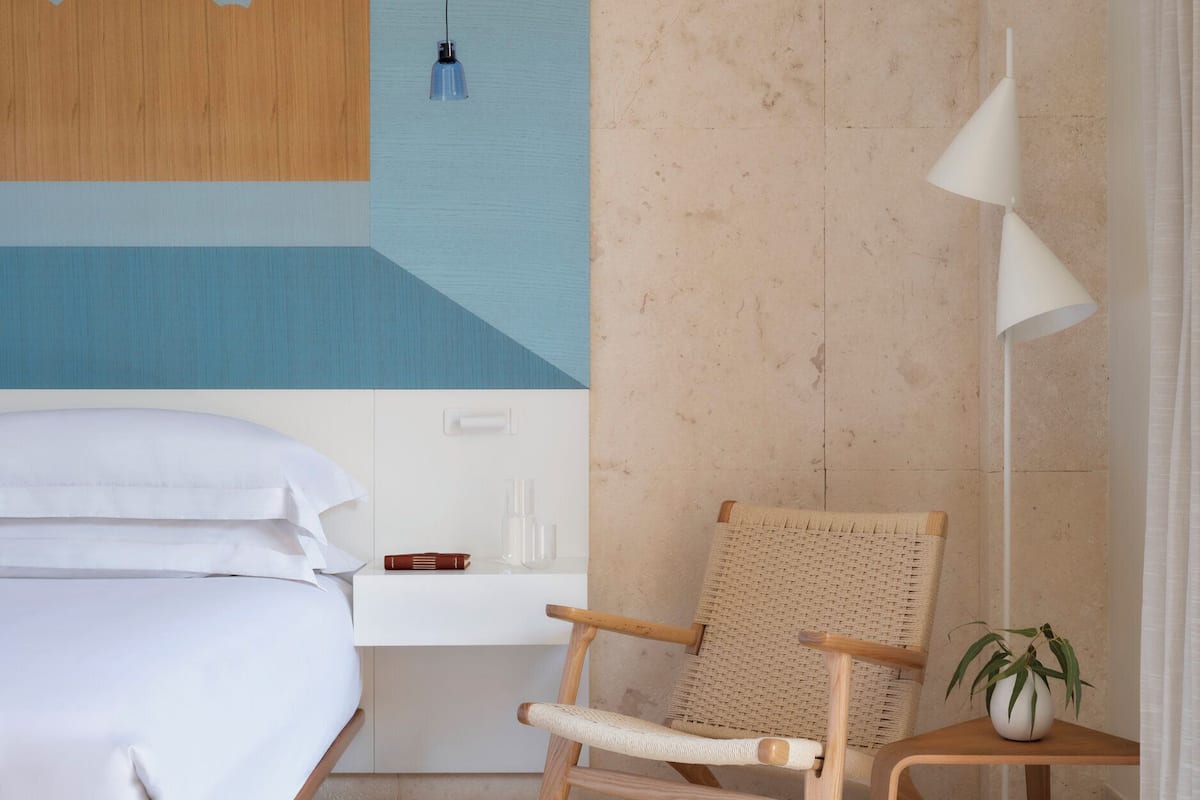 Ein Schlafzimmer mit Bett, Stuhl, Nachttisch, Lampe und einer Wand mit einem großen abstrakten Gemälde.