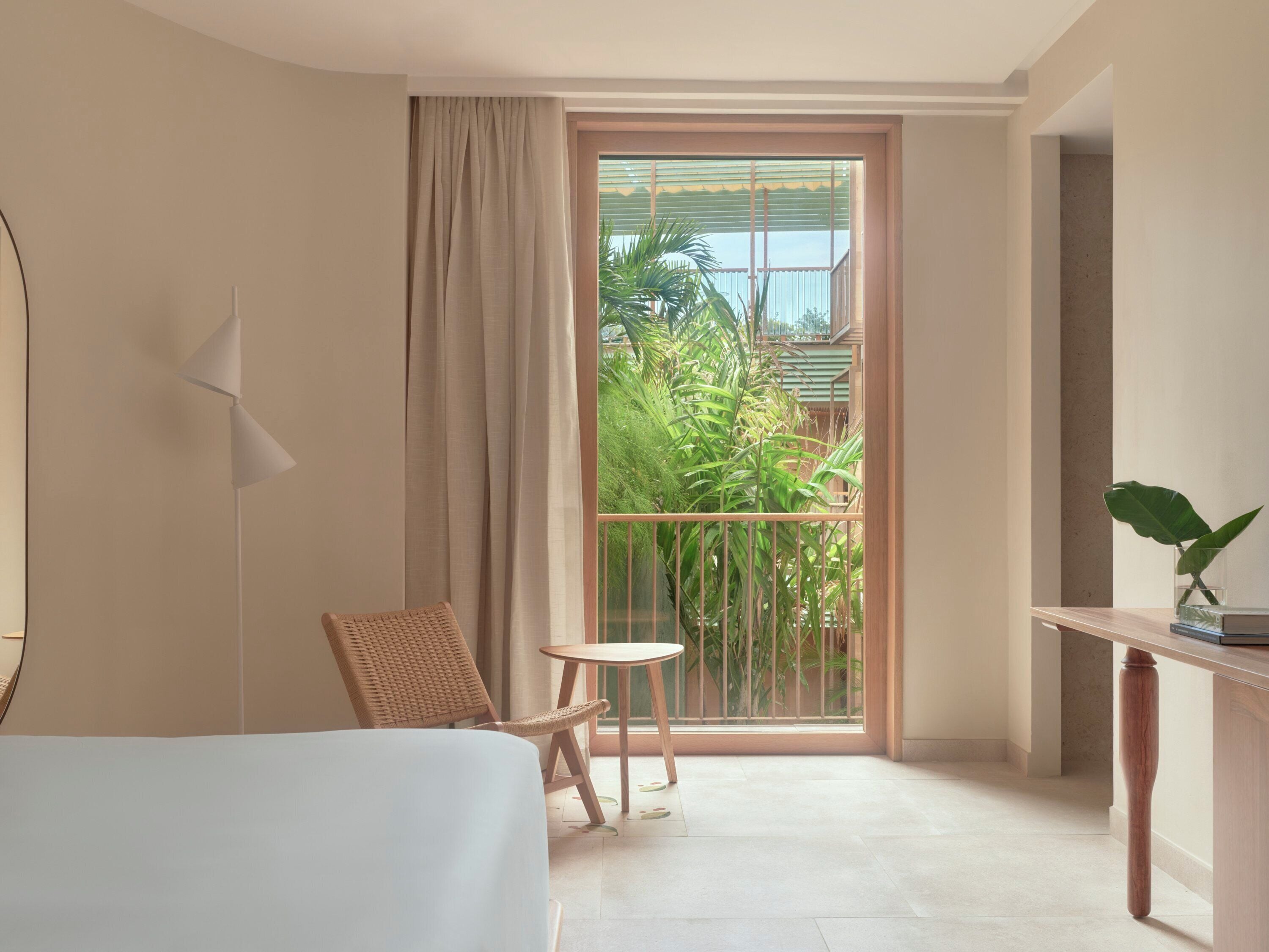 Quarto de hotel com cama, uma cadeira, uma mesinha e vista para o verde através de uma grande janela.