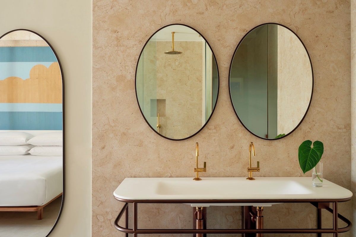 Ein Badezimmer mit zwei runden Spiegeln, einem weißen Waschbecken mit goldfarbenen Armaturen und einem Bett mit weißer Bettwäsche.