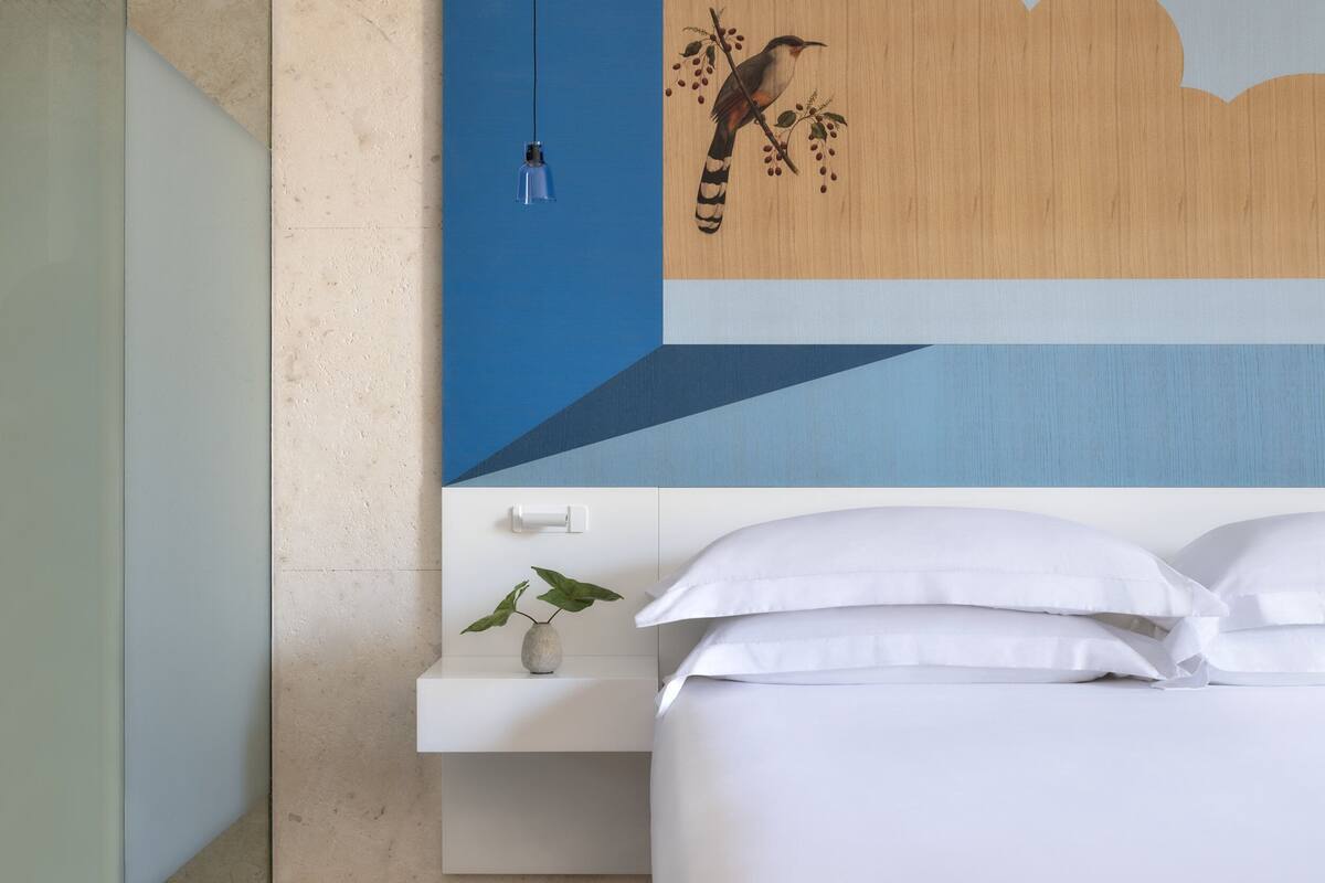 Ein modernes Hotelzimmer mit einem Bett, einem Nachttisch, einer Pflanze in einer Vase und einem Wandbild mit einem Vogel auf einem Ast.