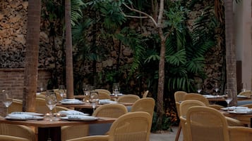 An outdoor dining area with wicker chairs and tables, surrounded by tropical plants and a stone wall.