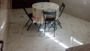 A dining area with a round table covered by a floral tablecloth, surrounded by four chairs. The room has a tiled floor and a window with blinds. - Belle Appartement Dans les Hauteurs de el Hajeb Proche de la Ville e Centre (El-Hajeb)