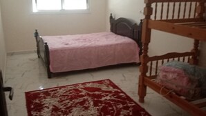A bedroom with a bunk bed, a single bed, a window, and a red and white patterned rug. - Belle Appartement Dans les Hauteurs de el Hajeb Proche de la Ville e Centre (El-Hajeb)