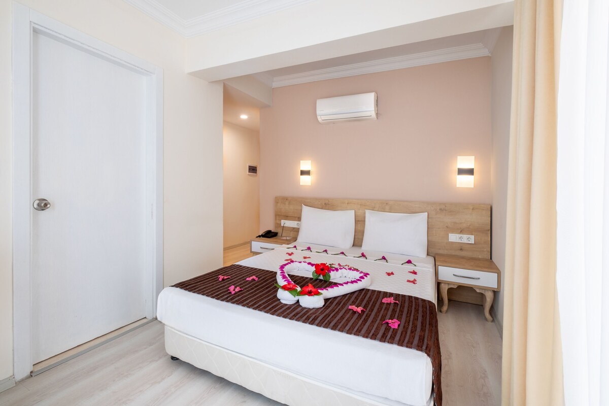 A hotel room with a bed, bedside tables, an air conditioning unit, and a door.