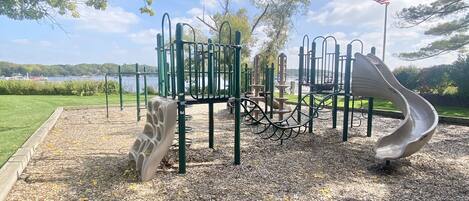 A playground with various equipment, including a slide and climbing structures, set on a gravel surface with trees in the background.