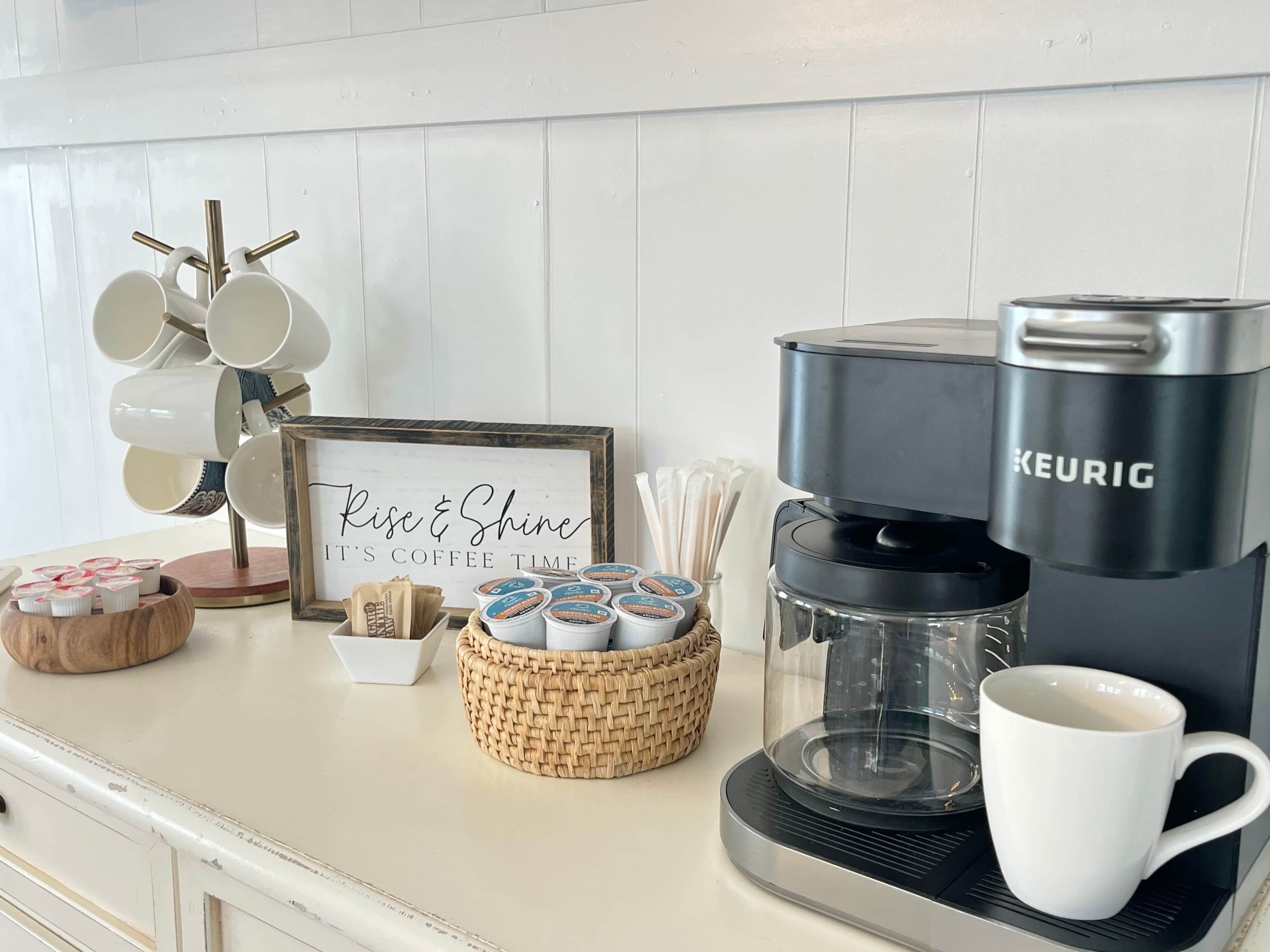 A coffee station with a Keurig machine, coffee pods, a mug, and a sign that says 'Rise & Shine It's Coffee Time.'