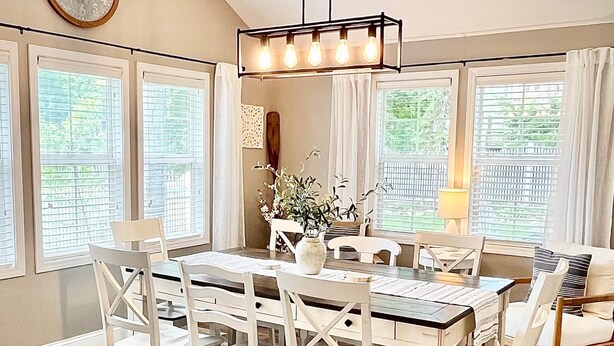 A dining area with a wooden table, white chairs, a hanging light fixture, and large windows with blinds. - Blue Cottage in the Bay - walk to beach and shops (Williams Bay)