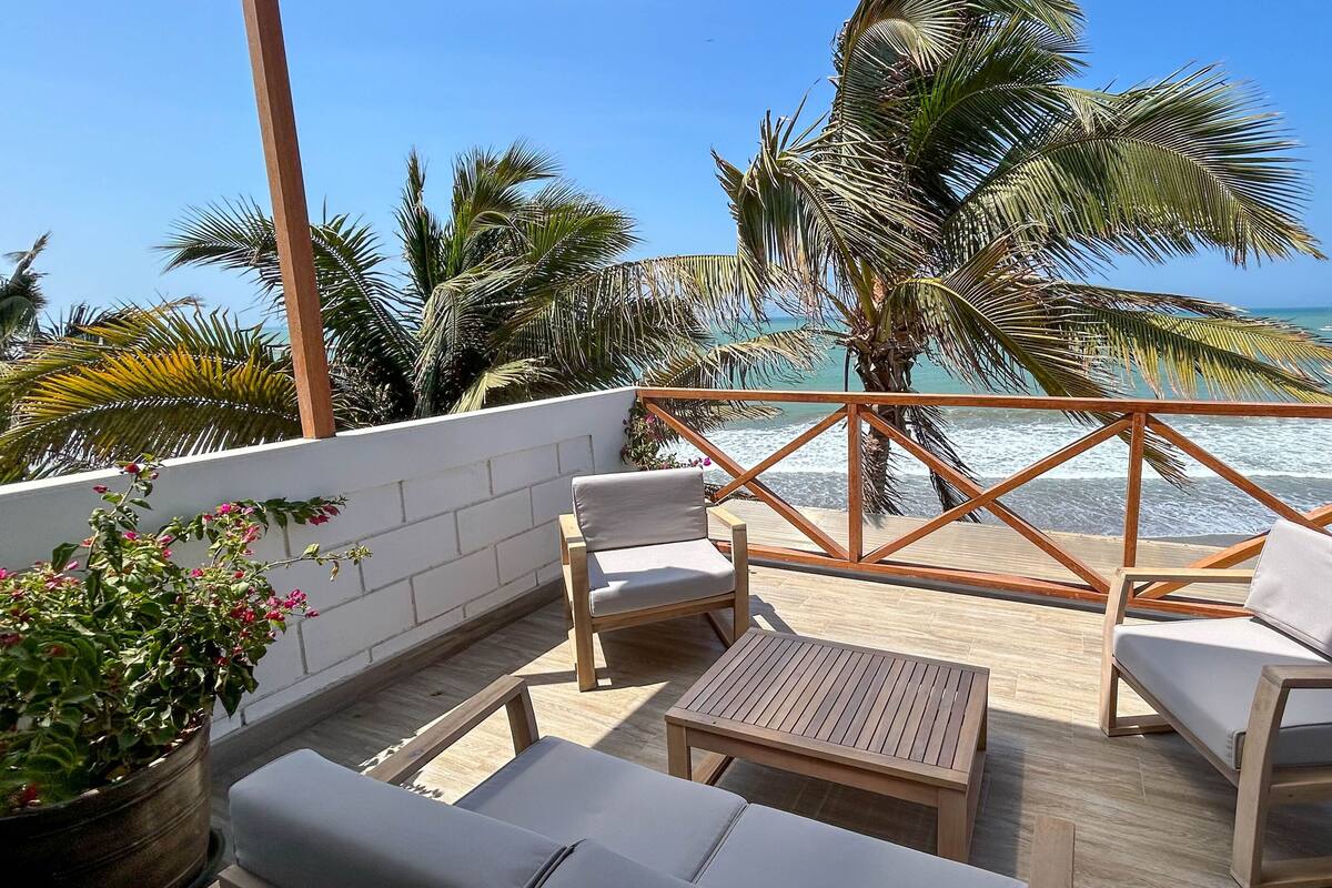 Una terraza con mobiliario de madera, vista al océano y palmeras.