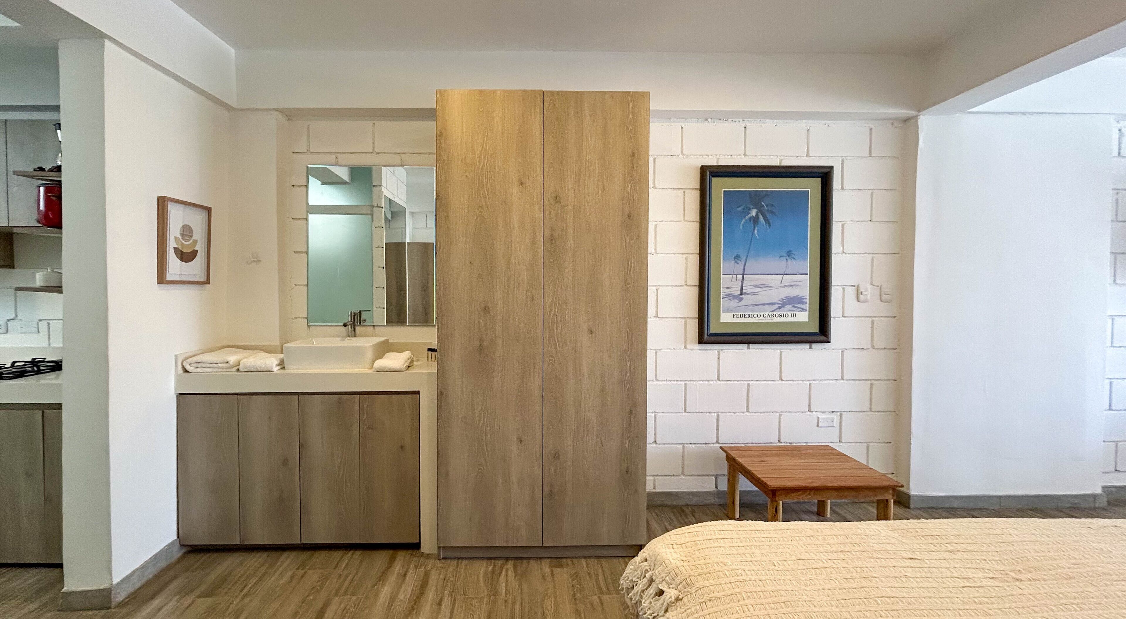 A bedroom with a wooden wardrobe, a bedside table, a framed picture on the wall, and a bathroom with a sink and mirror.
