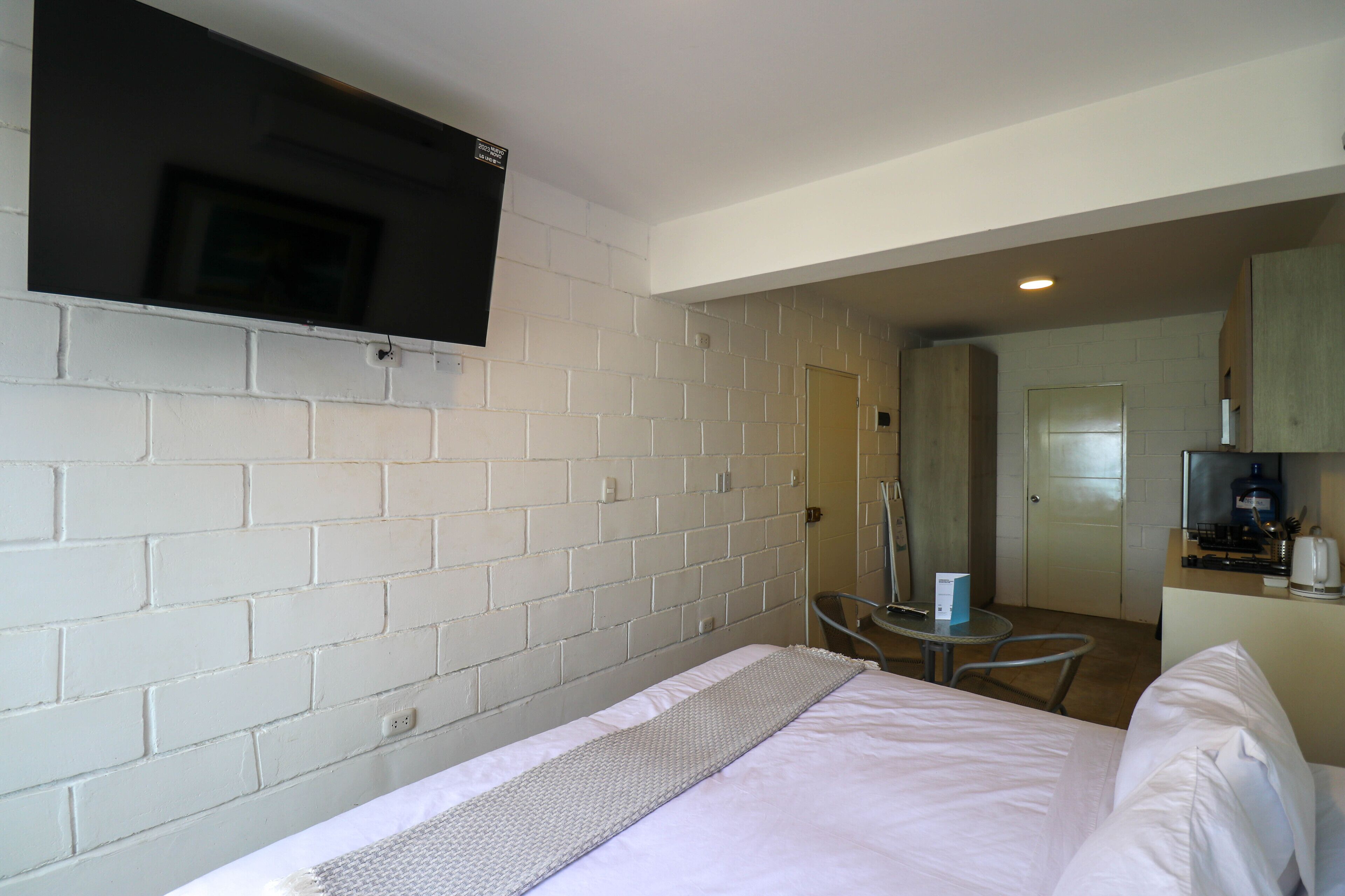 A hotel room with a bed, a TV mounted on the wall, a small table, and a kitchenette area.