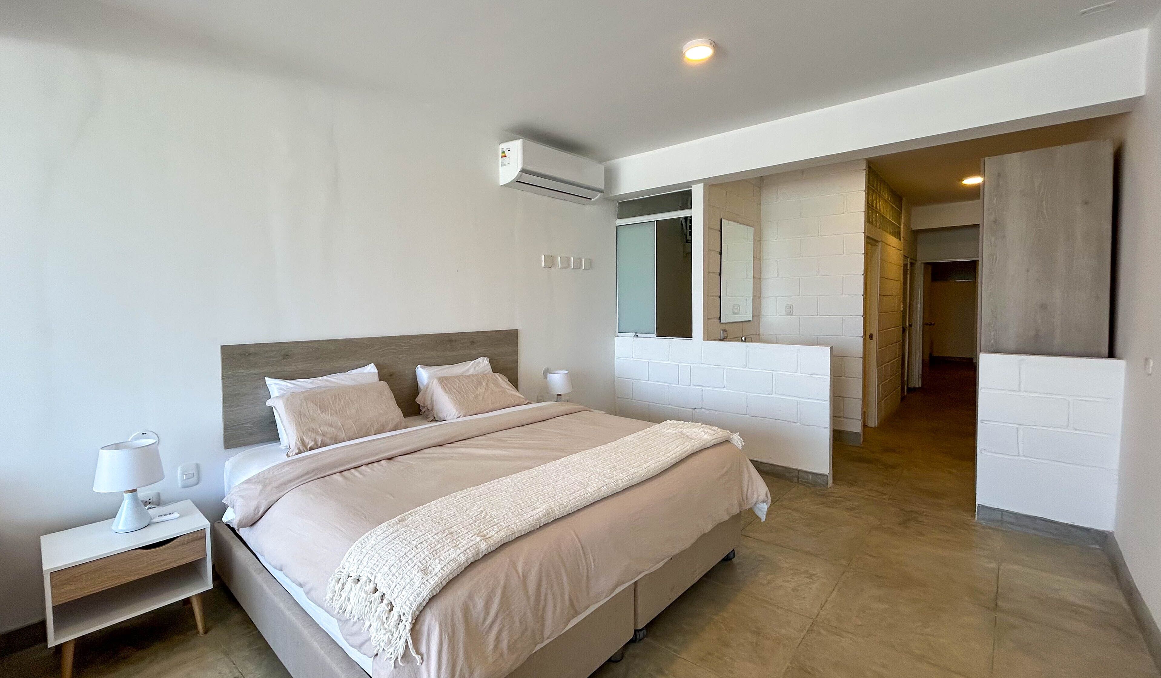 A modern bedroom with a large bed, a bedside table, a nightstand, a wall-mounted air conditioner, a mirror, and a bathroom visible through an open door.