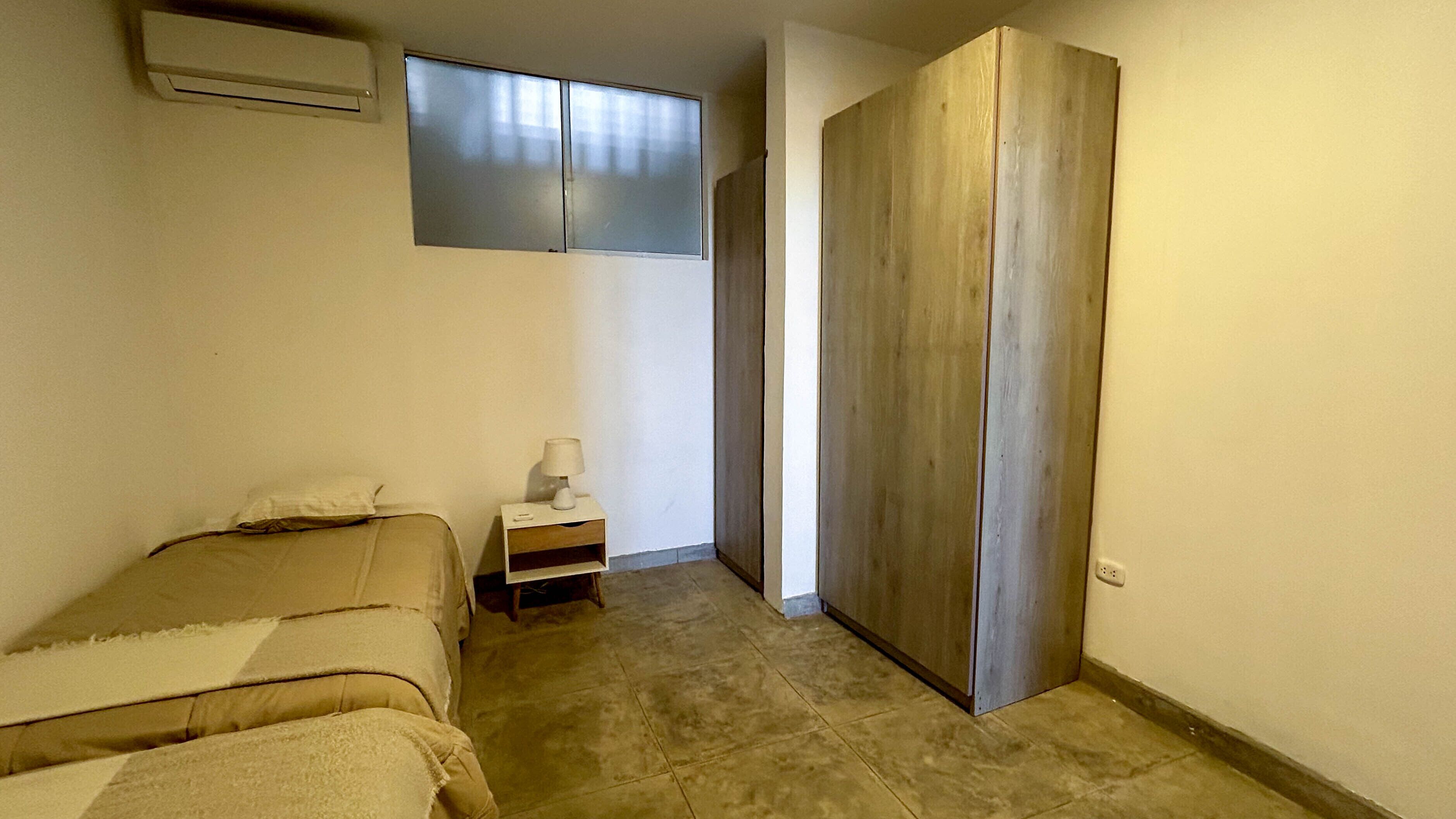A small, narrow room with a bed, a nightstand with a lamp, and a wardrobe.