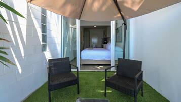 Un balcón con dos sillas de mimbre, un ventanal de vidrio que da a un dormitorio y un toldo grande que da sombra.