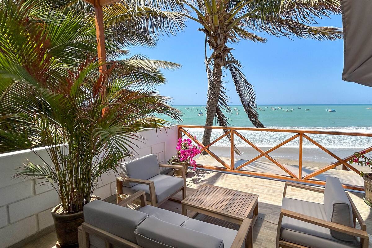 Un balcón con mobiliario de madera, plantas en macetas y vista a la playa y al océano.