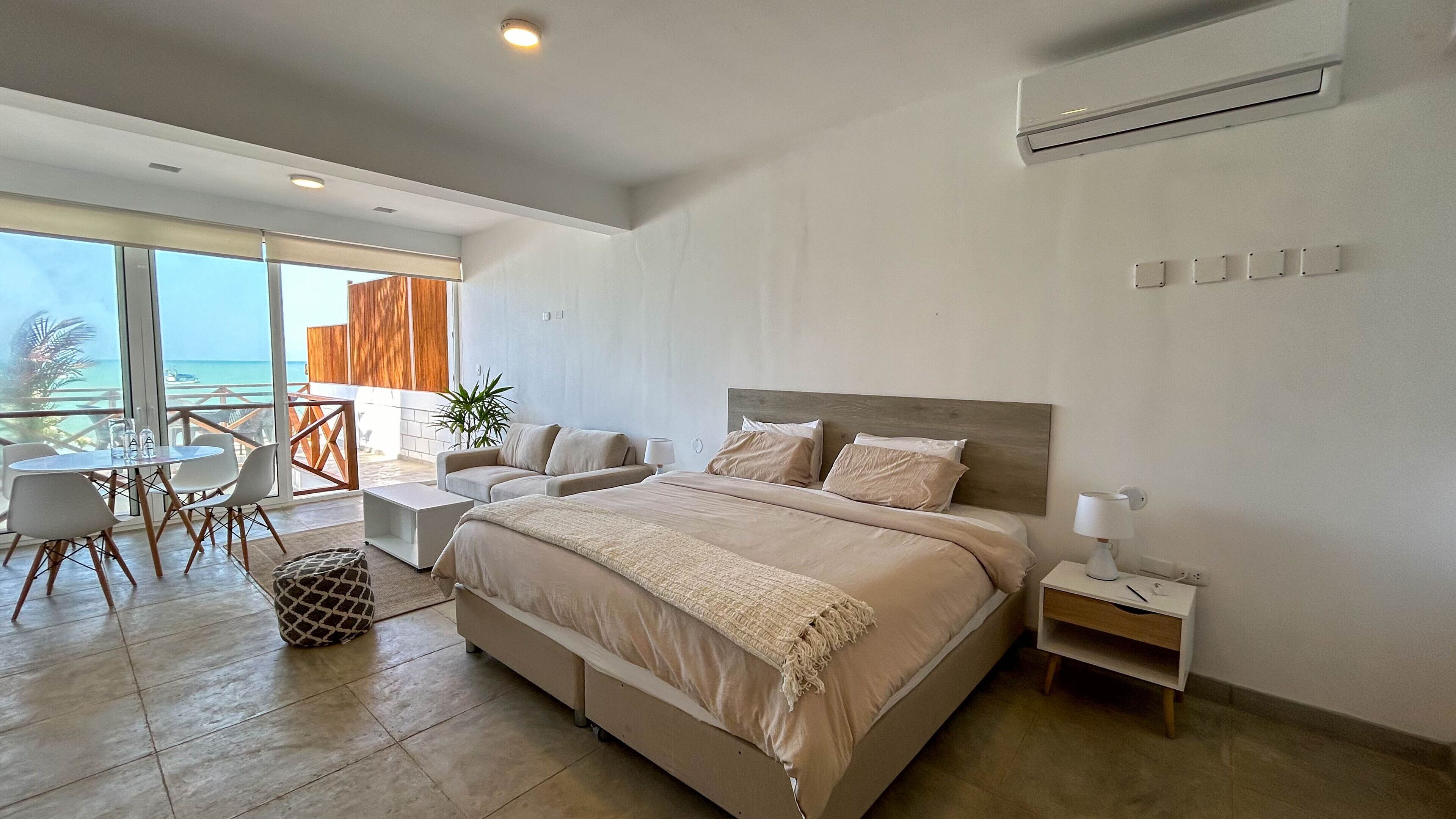A modern hotel room with a large bed, a balcony with a view of the sea, a dining area, and a small table with chairs.