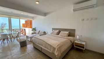 A modern hotel room with a large bed, a balcony with a view of the sea, a dining area, and a small table with chairs.