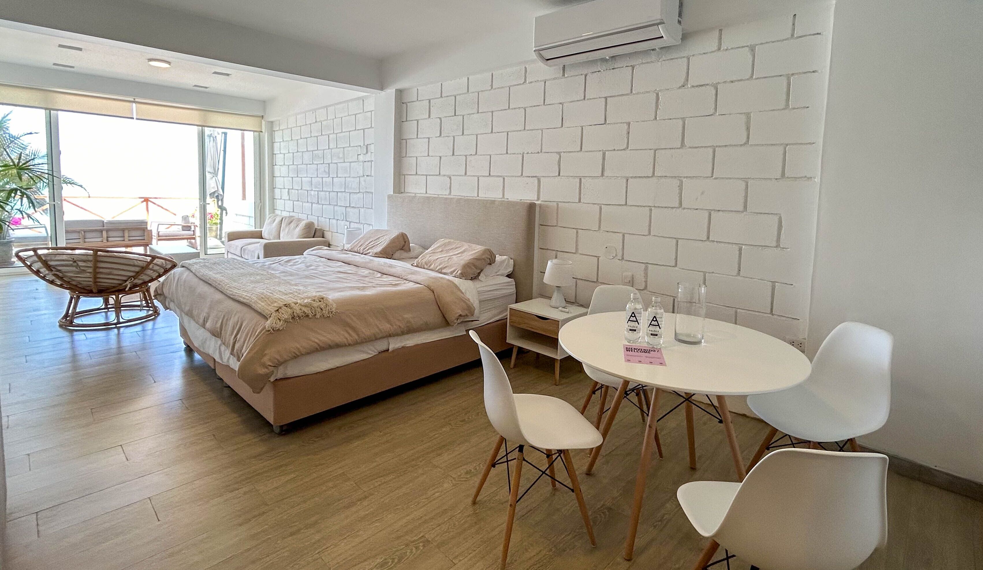 A modern hotel room with a large bed, a dining table with chairs, a small bedside table, and a white brick wall.