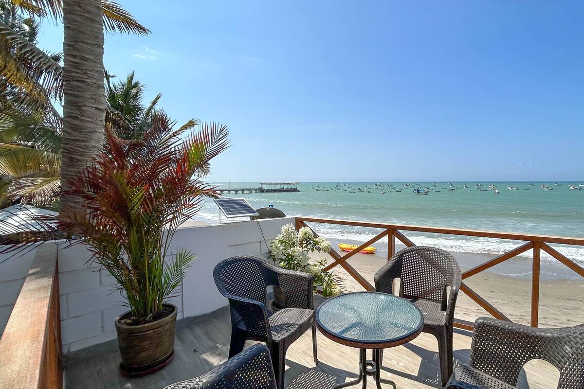 Un patio frente a la playa con muebles de mimbre y una mesa de vidrio, con vista al mar, barcos y un muelle a lo lejos.