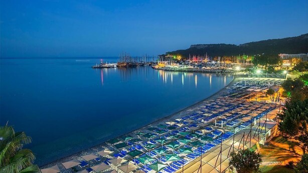 Un hotel frente al mar con zona de playa que incluye hileras de sombrillas y tumbonas, un puerto deportivo con barcos, y un acantilado con vistas al mar.