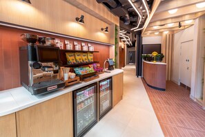 A coffee shop interior with a coffee machine, a display of coffee pods, a refrigerated beverage cooler, and a counter with a cash register. - YOTELPAD London Stratford (London)