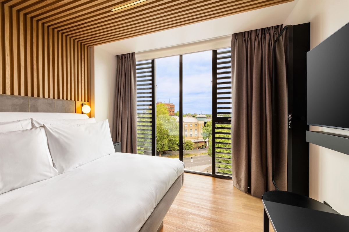 Une chambre d’hôtel avec un lit, une télévision, une fenêtre avec des rideaux et une vue sur les bâtiments environnants.
