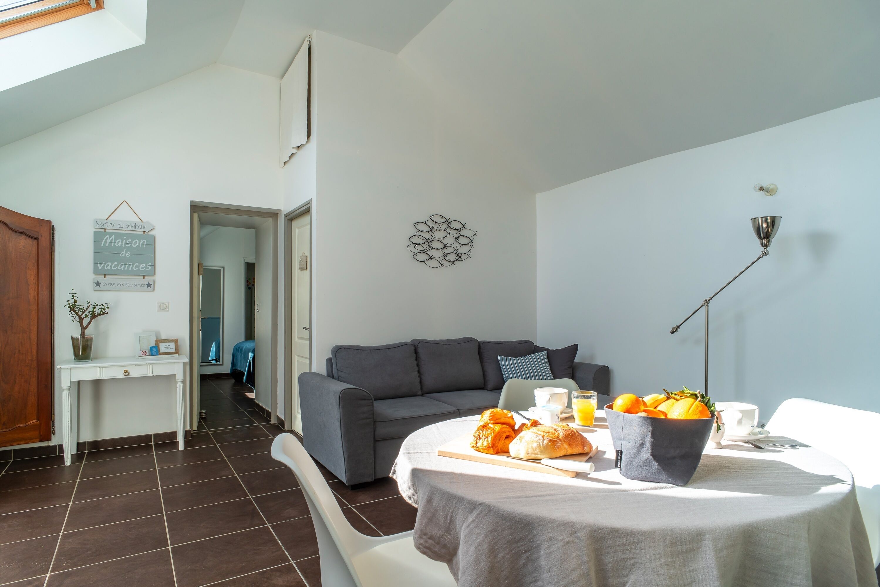 A modern living room with a grey sofa, a dining table with breakfast items, and a white wall with a decorative piece.