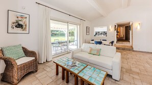 A spacious living area with a white sofa, a wicker chair, and a coffee table. There is a large sliding glass door leading to an outdoor patio. - Villa Triptyque - Avec piscine et grand jardin (Villecroze)