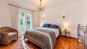 A bedroom with a bed, a wicker chair, a chandelier, a lamp, and a view of the outdoors. - Villa Triptyque - Avec piscine et grand jardin (Villecroze)