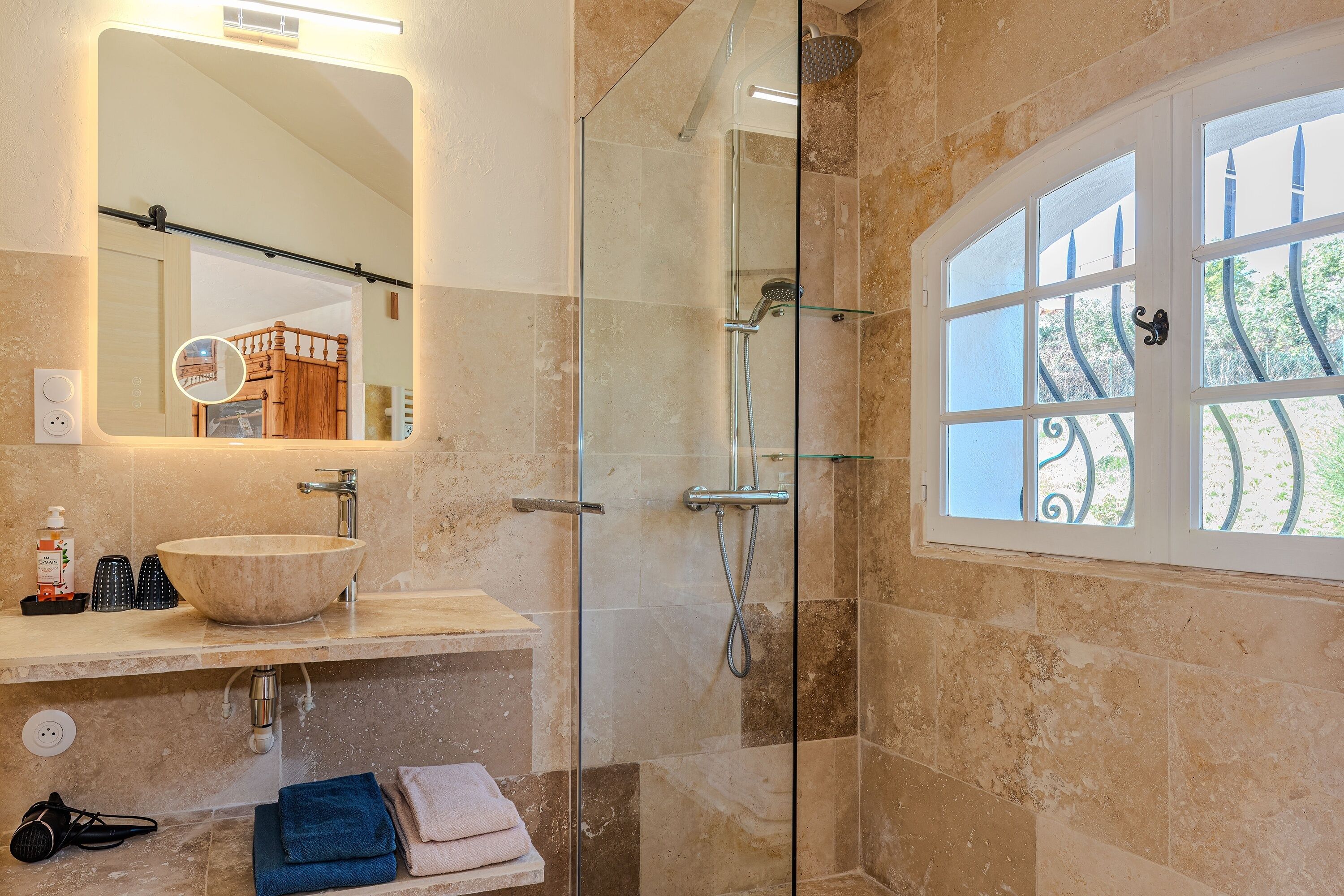 Une salle de bain dotée d’une douche à parois de verre, d’un grand miroir, d’un lavabo avec un receveur et d’une fenêtre ornée de barreaux décoratifs.