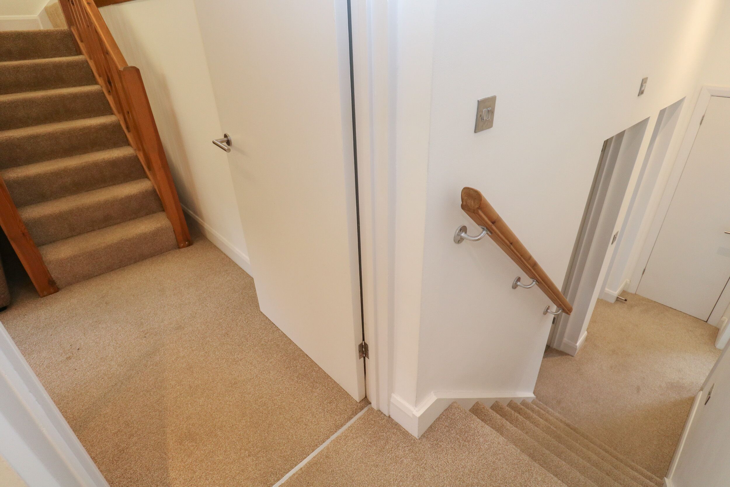 A carpeted staircase with wooden handrails, white walls, and a door leading to another room.