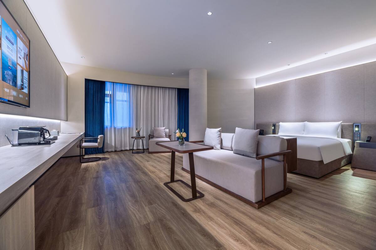 Une chambre d’hôtel moderne avec un plancher en bois, un canapé, un lit, un bureau avec un ordinateur portable et un téléviseur fixé au mur.