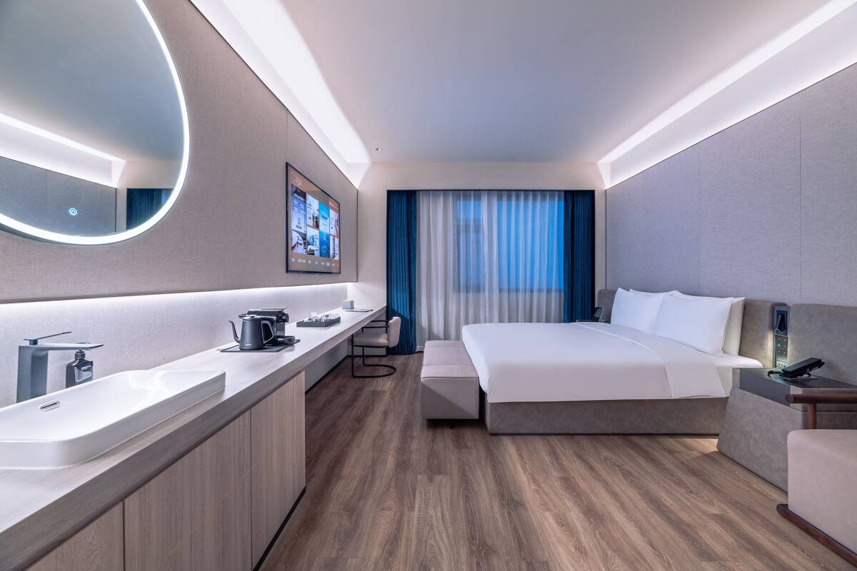 Une chambre d’hôtel moderne avec un grand lit, une table de chevet, un bureau avec une chaise, un grand miroir et un téléviseur fixé au mur.