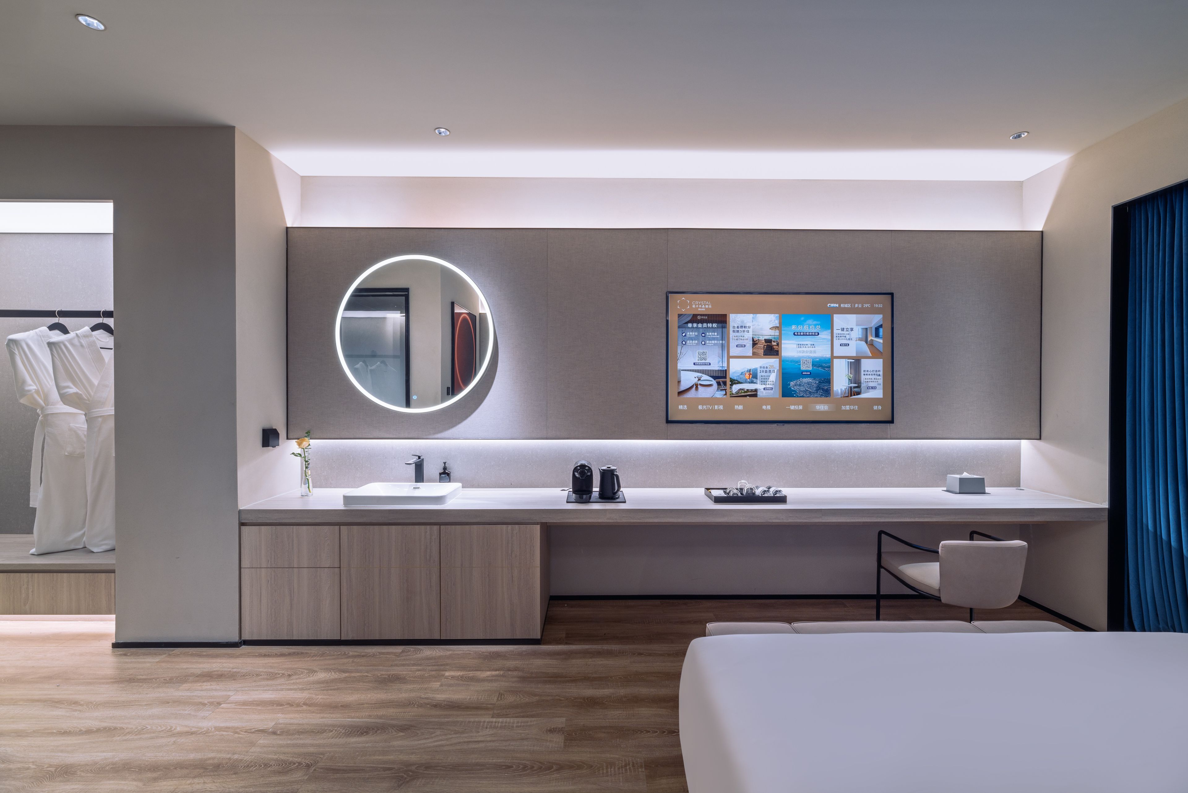 A modern hotel room with a large bed, a built-in desk with a computer, a round mirror, and a wall-mounted TV displaying room information.