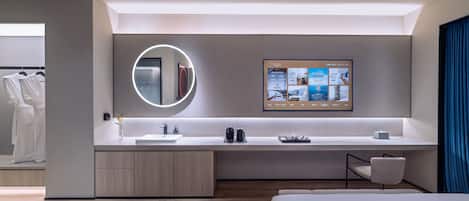 A modern hotel room with a large bed, a built-in desk with a computer, a round mirror, and a wall-mounted TV displaying room information.