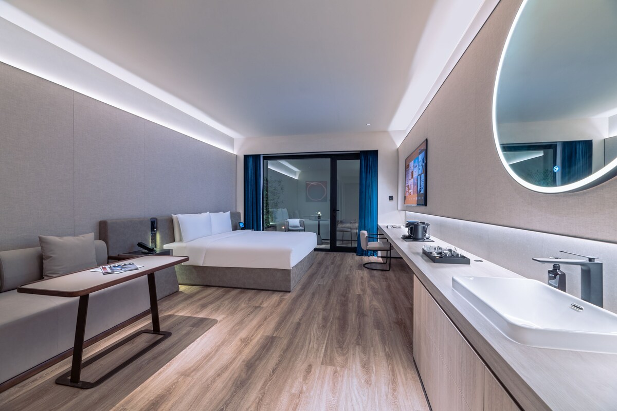 Une chambre d’hôtel moderne comprenant un lit, un canapé, une petite table, un bureau, une salle de bain avec un lavabo et un miroir, et une vue sur l’extérieur grâce à une porte vitrée.