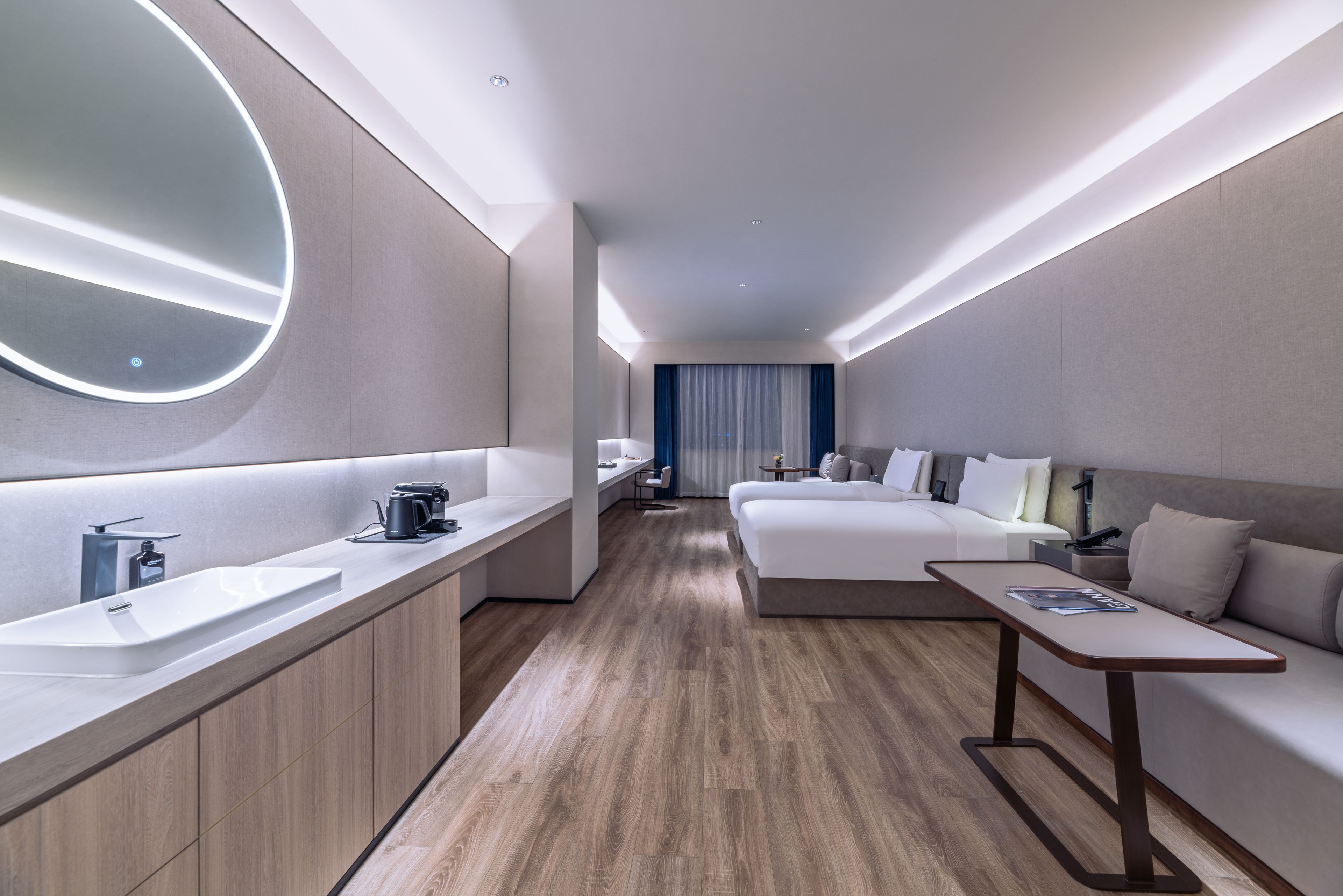A modern hotel room with a large bed, a bedside table, a desk, a round mirror, and a bathroom with a sink and toilet.
