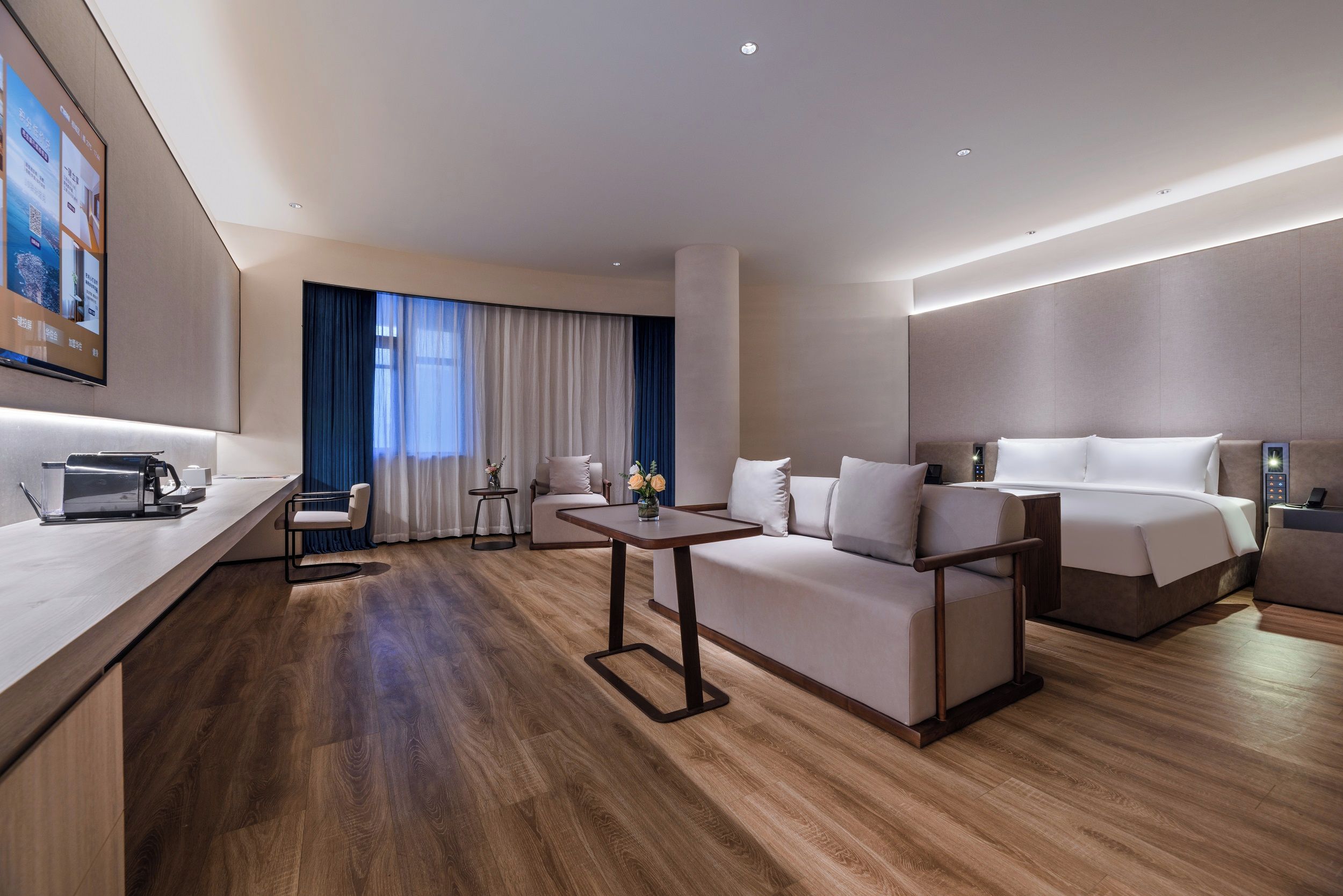 Quarto de hotel moderno com piso de madeira, sofá, cama, mesa pequena e televisão na parede.