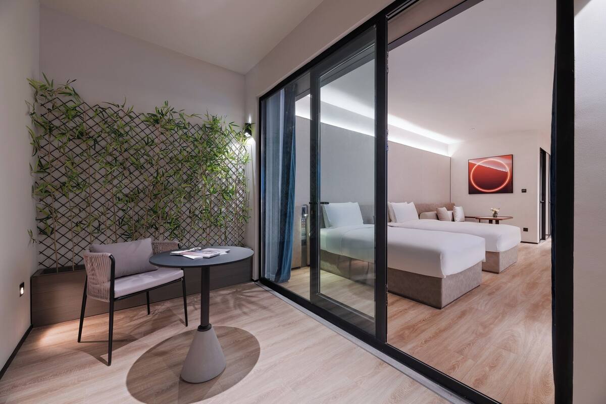 Une chambre d’hôtel moderne dotée d’une porte-fenêtre coulissante donnant sur une chambre à coucher, d’un coin salon avec un canapé et une table, et d’un mur décoratif en bambou.