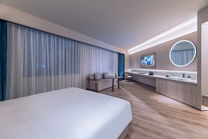 Moderni hotellihuone, jossa on suuri sänky, sohva, työpöytä, pyöreä peili ja seinälle asennettu televisio.