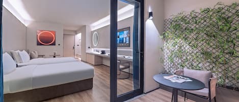 A modern hotel room with a large bed, a small table, a chair, a TV, a mirror, and a wall with a bamboo plant.