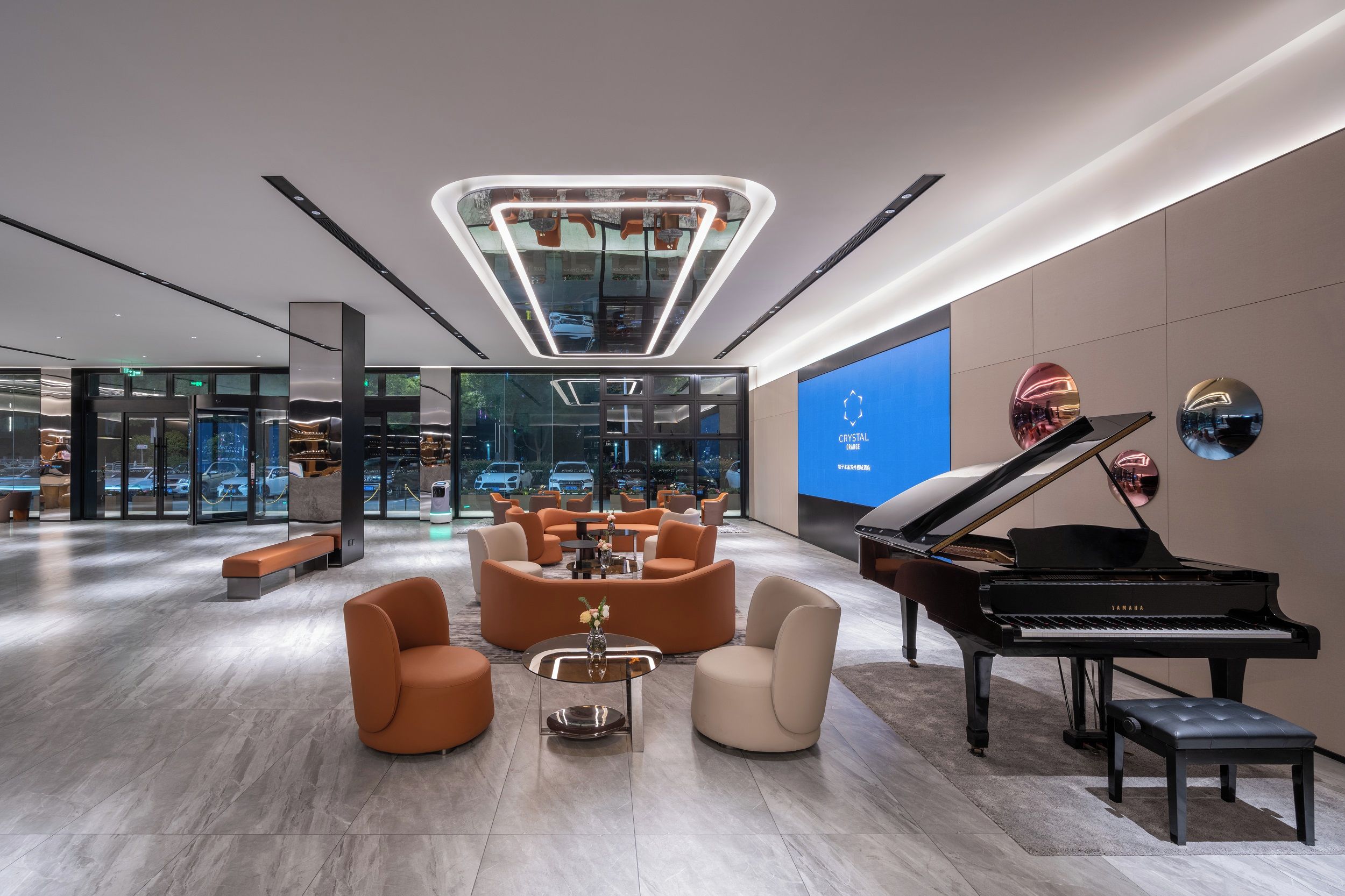 A modern lobby with a grand piano, comfortable seating, a central light fixture, and a large digital screen displaying the brand name.